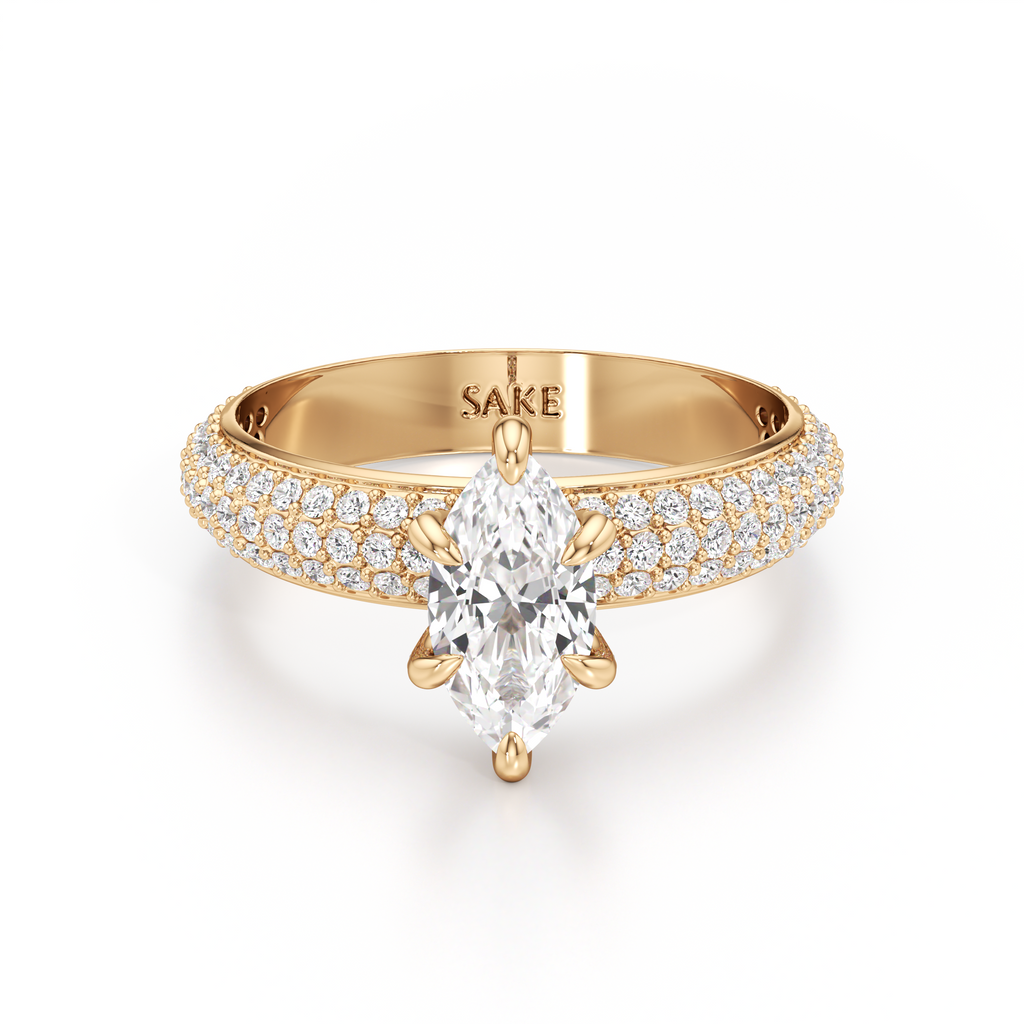 A yellow gold engagement ring featuring a marquise-cut clear center stone and pavé-set diamonds along the shoulders, shown on a clean white background. The inside of the band is engraved with the brand name 'SAKE'.