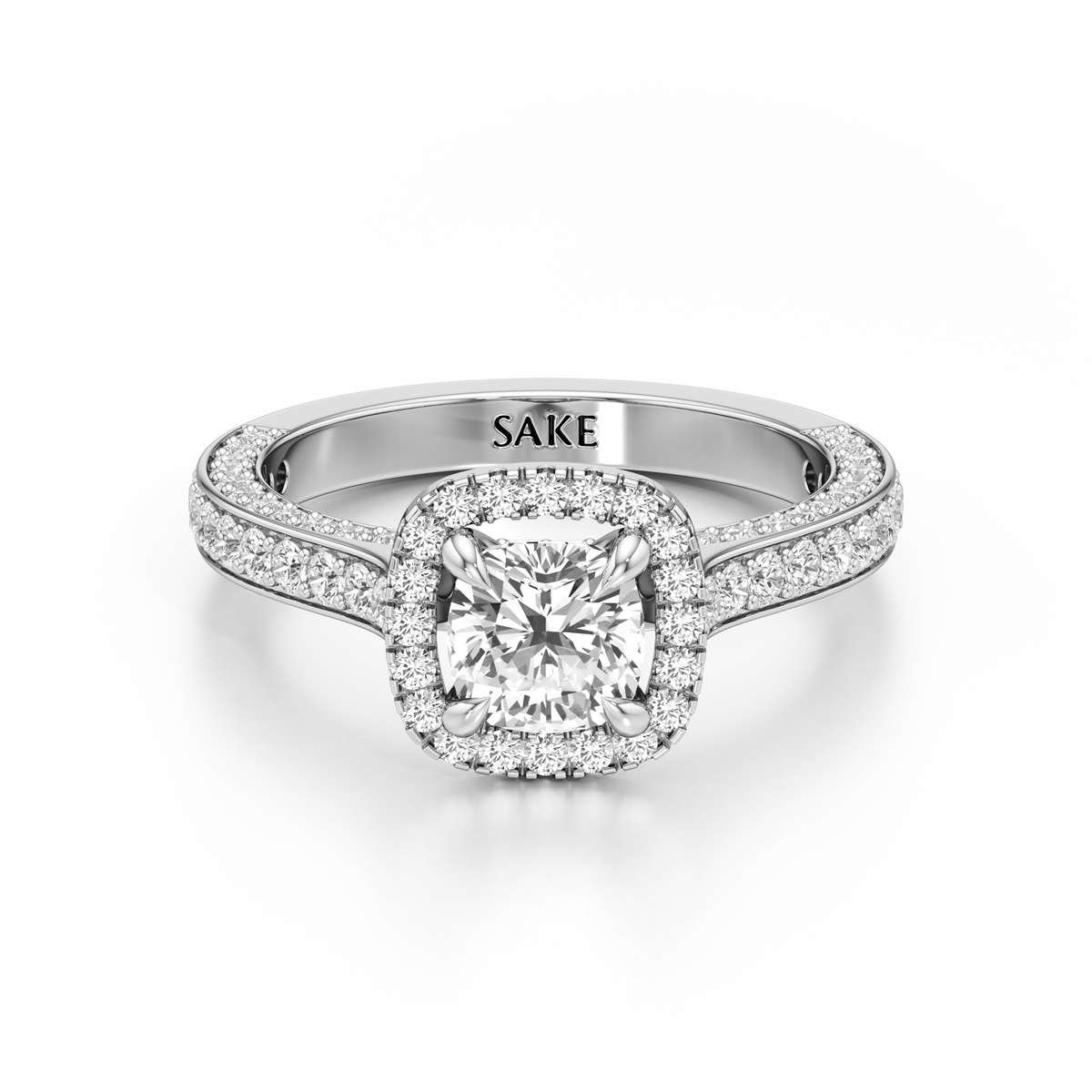 A silver-toned halo engagement ring with a cushion-cut center diamond and pavé-set side stones, shown on a clean white background. The inside of the band is engraved with the brand name 'SAKE'.