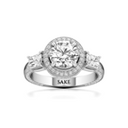 A silver-toned diamond ring with a round-cut center stone, a halo of small diamonds, and three marquise-cut side stones. The band is engraved with the name 'SAKE'.