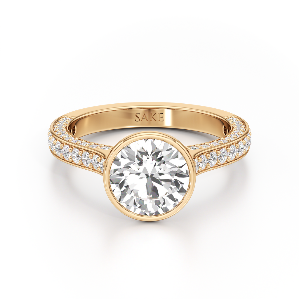 A yellow gold engagement ring featuring a large round-cut center stone and pavé-set side stones on a clean white background. The inside of the band is engraved with the brand name 'SAKE'.