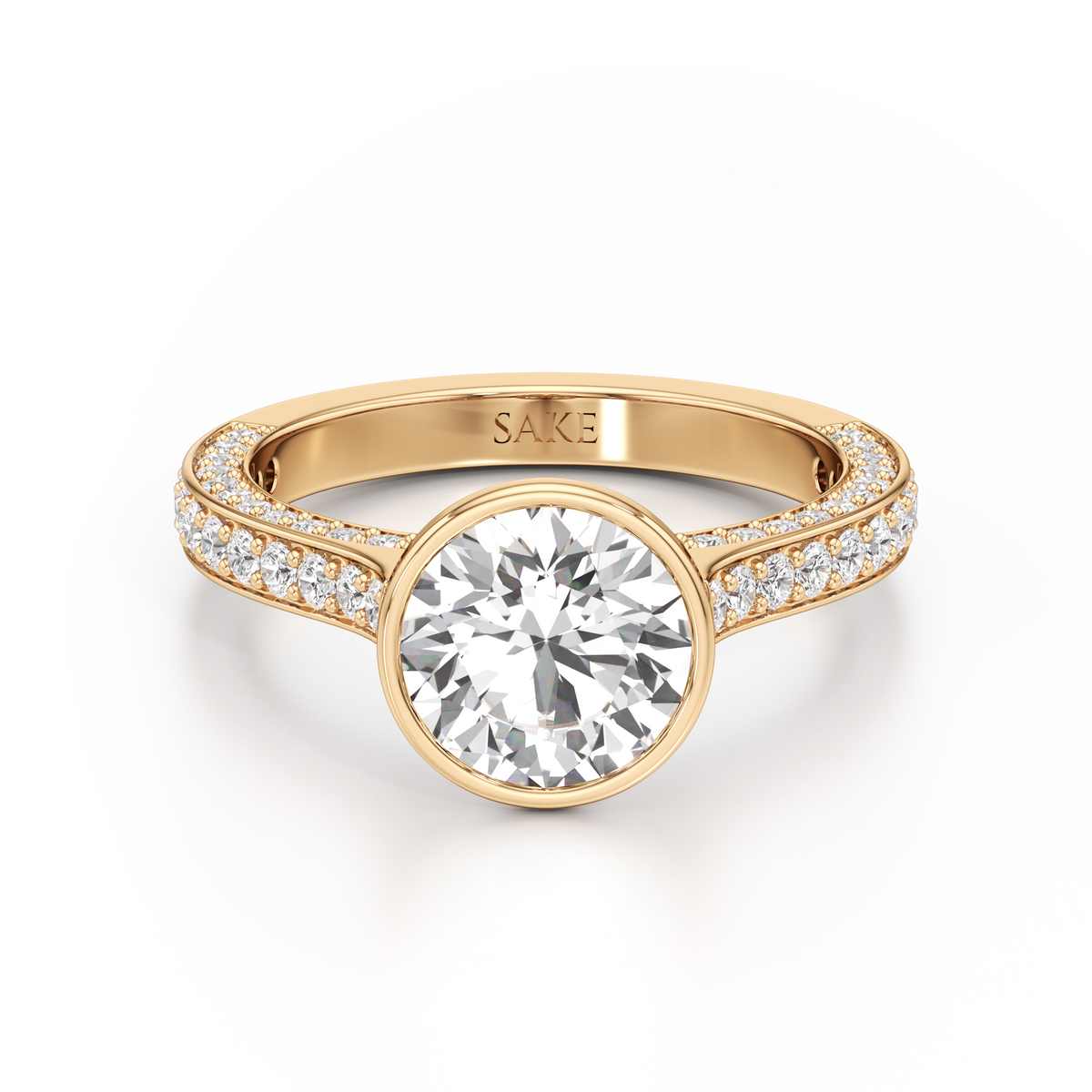A yellow gold engagement ring featuring a large round-cut center stone and pavé-set side stones on a clean white background. The inside of the band is engraved with the brand name 'SAKE'.