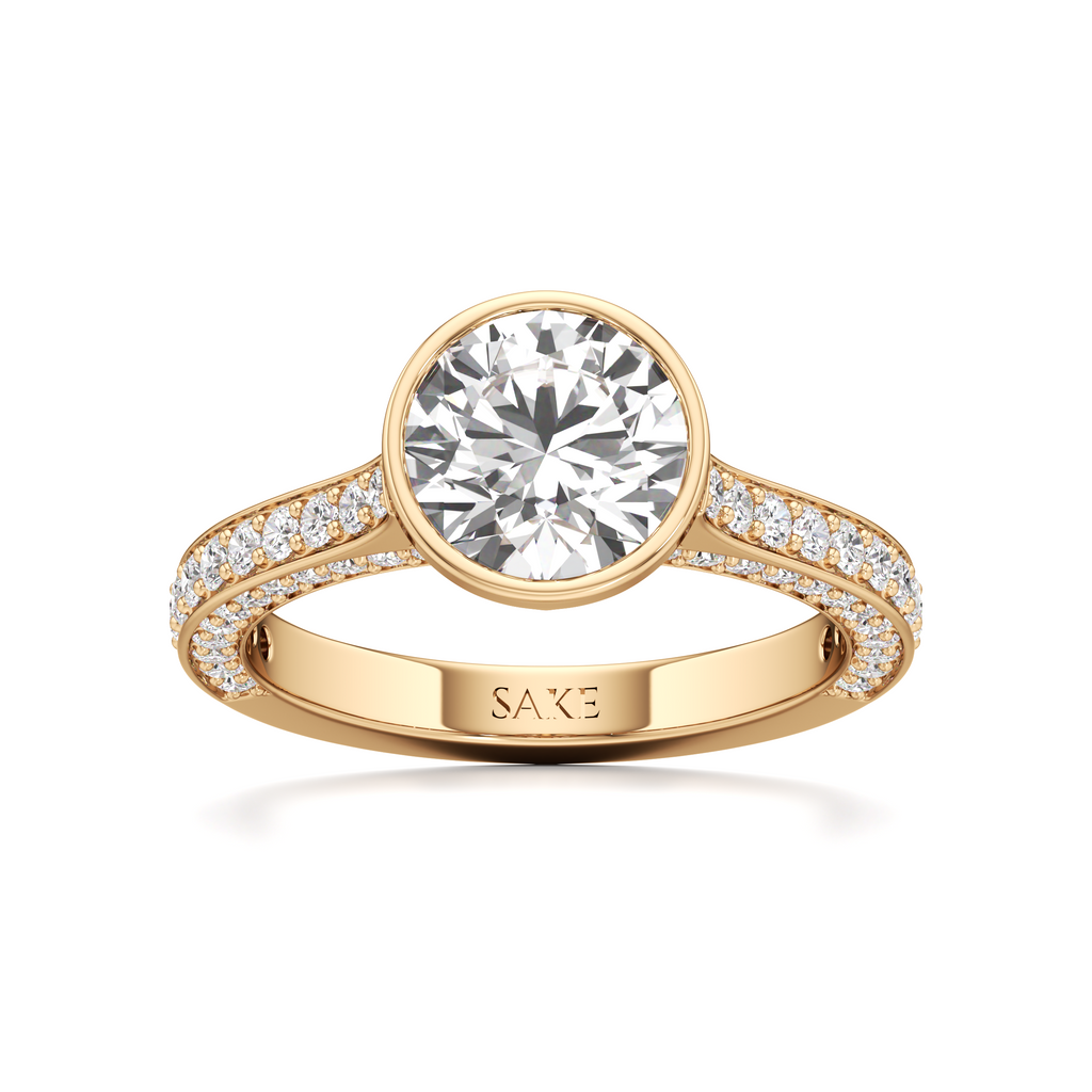 A yellow gold solitaire engagement ring featuring a round brilliant-cut diamond center stone and a pavé-set diamond band, with the brand name 'SAKE' engraved on the inner band, set against a clean white background.