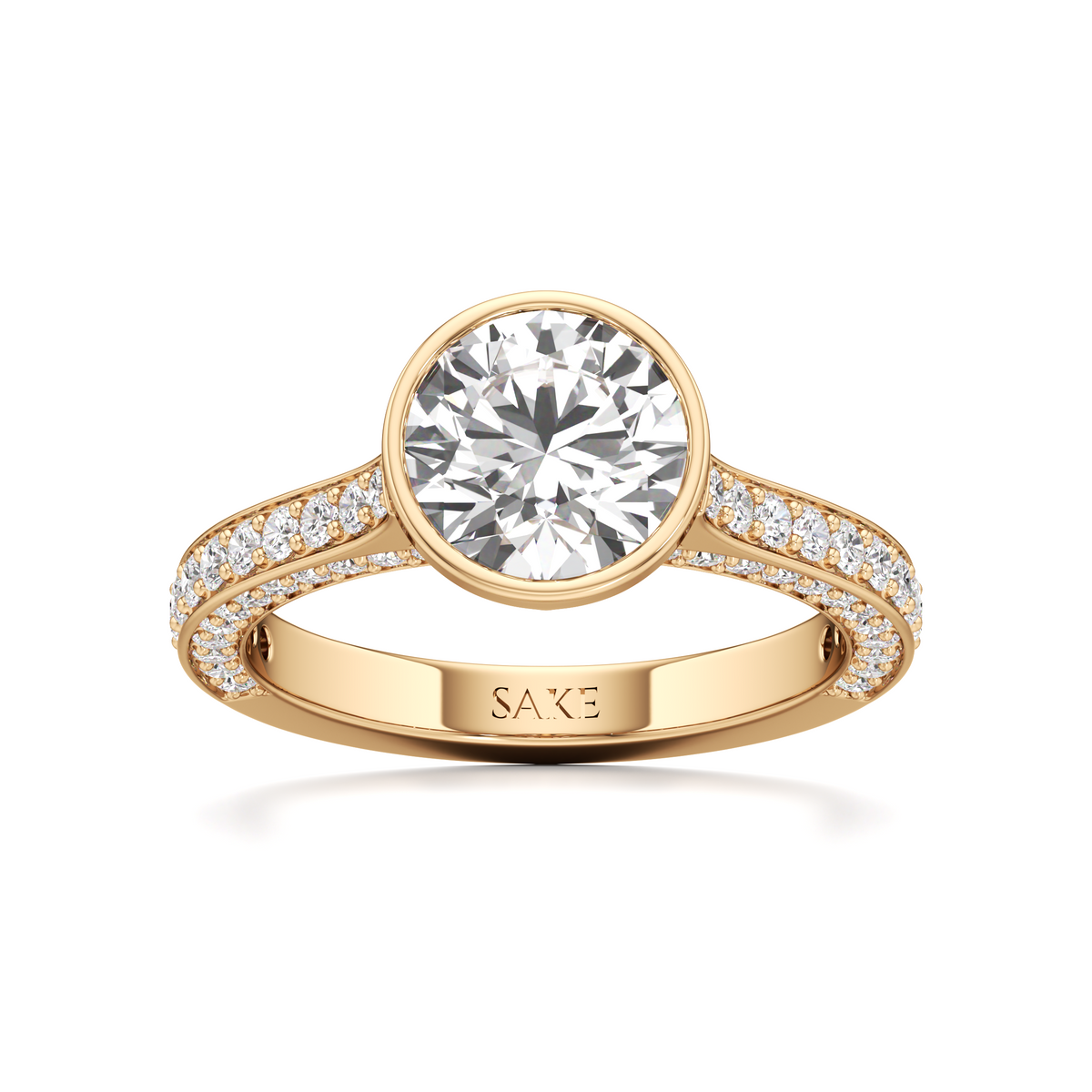 A yellow gold solitaire engagement ring featuring a round brilliant-cut diamond center stone and a pavé-set diamond band, with the brand name 'SAKE' engraved on the inner band, set against a clean white background.