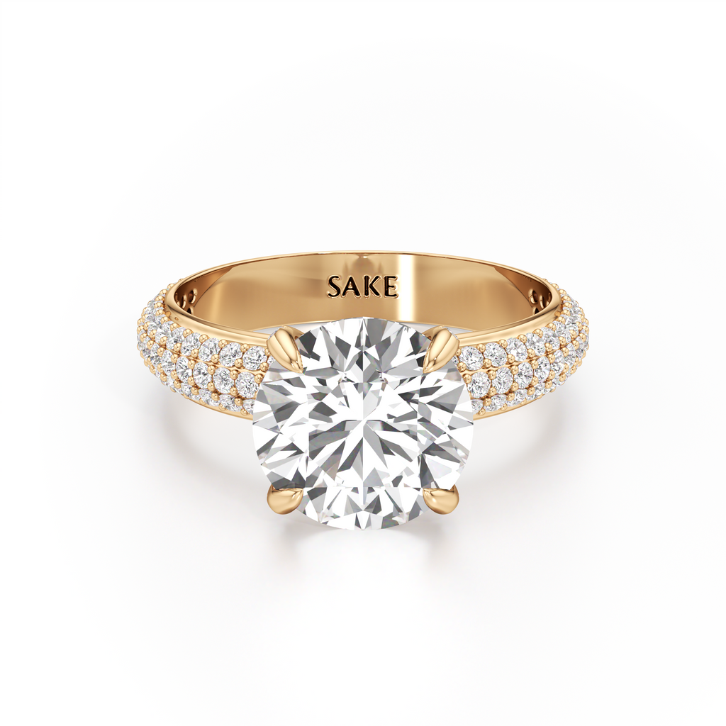 A yellow gold engagement ring featuring a large round-cut center diamond and pavé-set diamonds along the shoulders, with the brand name 'SAKE' engraved inside the band, shown on a clean white background.