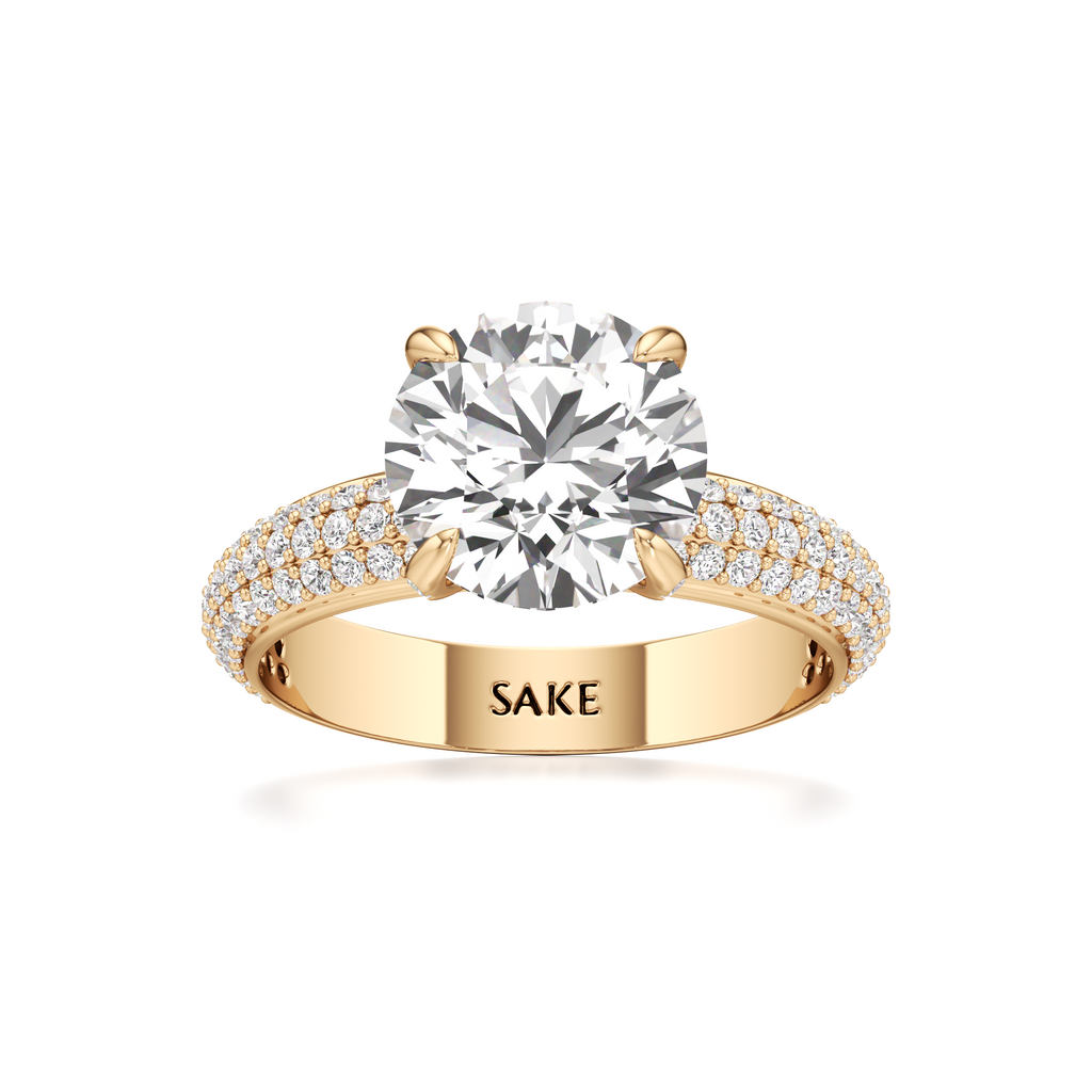 A yellow gold solitaire engagement ring with a round-cut diamond center stone and pavé-set diamonds on the band, featuring the engraved brand name 'SAKE' on the inside of the band.
