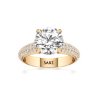 A yellow gold solitaire engagement ring with a round-cut diamond center stone and pavé-set diamonds on the band, featuring the engraved brand name 'SAKE' on the inside of the band.